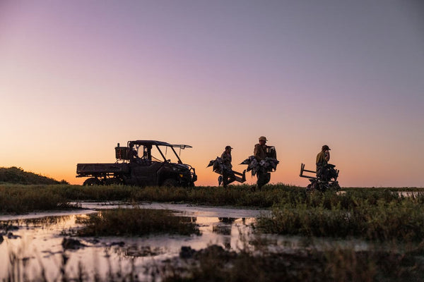 Three people on all-terrain vehicles in a field at sunset.