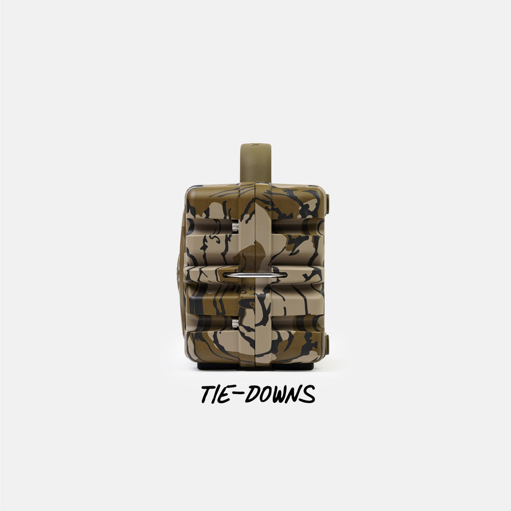 A tactical backpack in a camouflage pattern of tan, brown, and black, shown from the back with a metal ring rack and a strap tied down, on a light gray background with the text 'TIE-DOWNS' underneath.