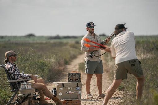 Two men in a field with one holding a bow and another sitting on a chair with a cooler.