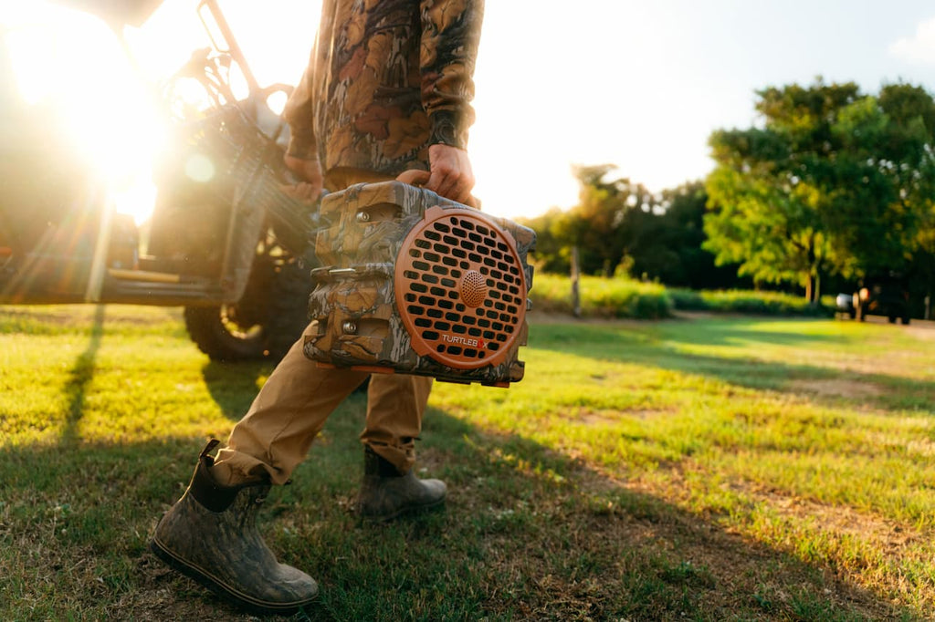 A person outdoors in a grassy field at sunset carrying a camouflage-patterned portable speaker with a prominent orange front grille.