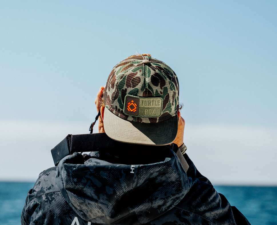 Lifestyle photo of a man facing the ocean with his back to the camera, wearing the All-Mesh Camo trucker hat