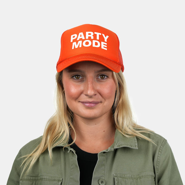 A lifestyle photo of a woman wearing a bright orange baseball cap with 'PARTY MODE' printed in white capital letters on the front, paired with an olive green shirt against a light gray background.