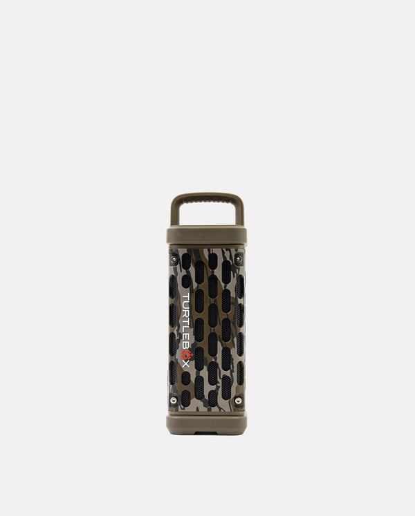 A portable cylindrical speaker with a camouflage pattern in olive green, brown, and black, featuring a top carrying handle and the Turtles X logo printed vertically on the side.