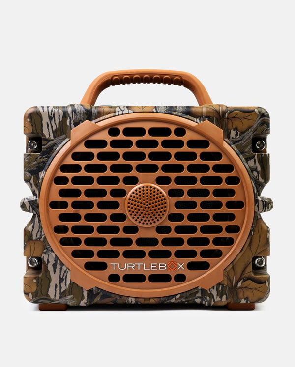 A rugged portable speaker with a camouflage pattern body, a brown handle, and a large circular brown front grille. The brand name 'TURTLEBOX' is printed on the front.