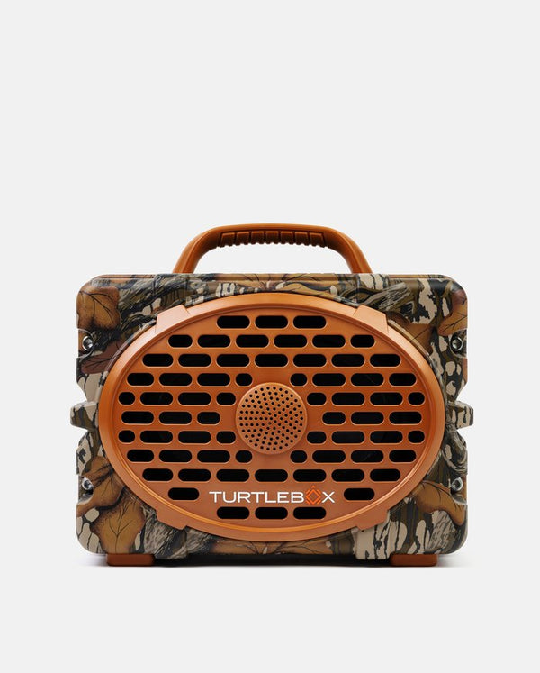 A Turtle X portable speaker with a brown camouflage pattern body, a prominent orange-brown oval front grille, and a matching handle on top, shown against a plain light gray background.