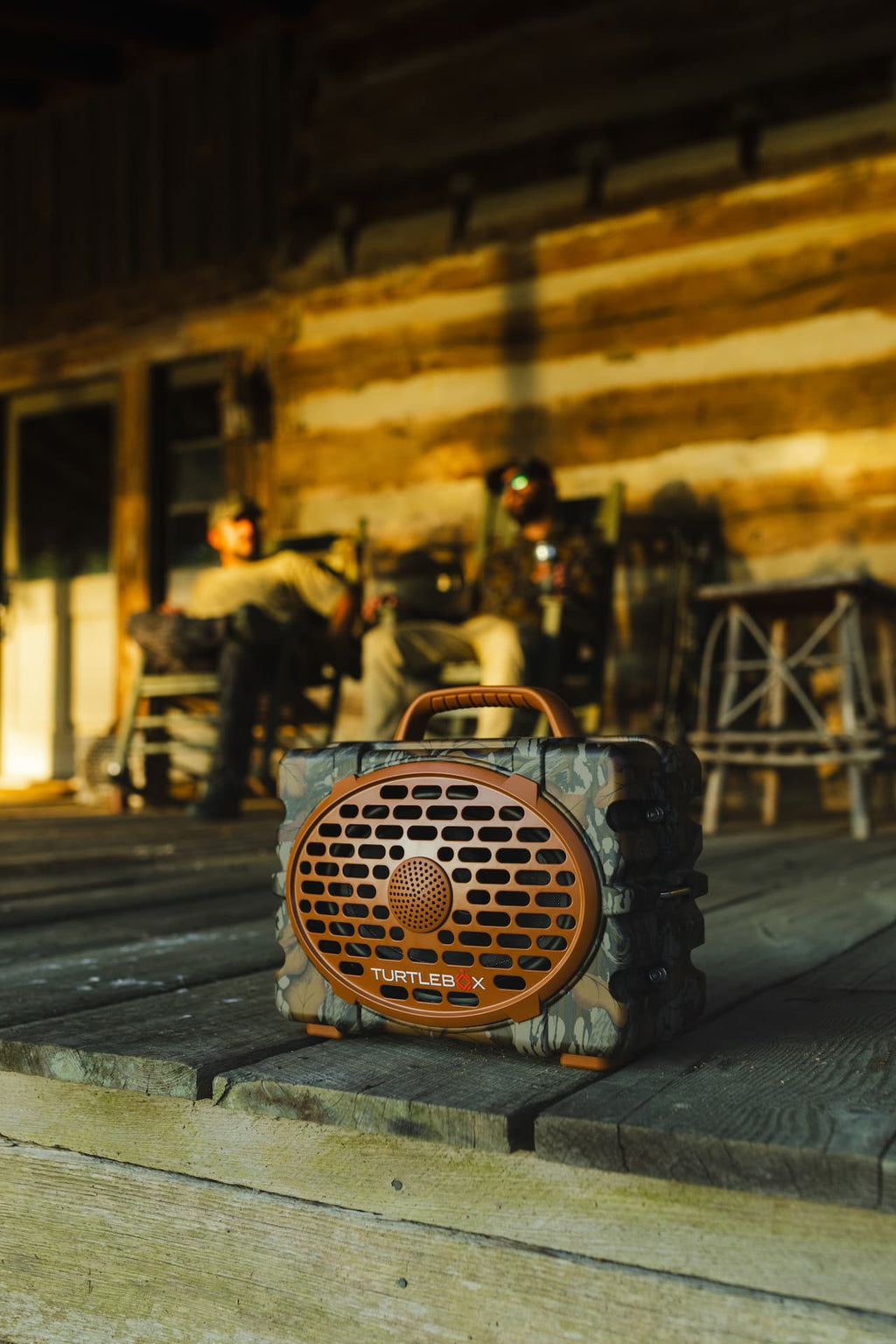 A rugged portable speaker with a camouflage pattern and copper-colored front grille sits on a wooden porch in front of a log cabin, with two people blurred in the background.