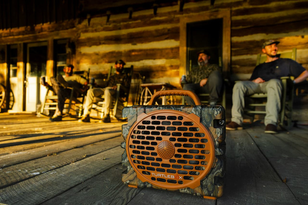A Catler X portable speaker with a camouflage pattern and orange front grille sits on a rustic wooden porch in front of a log cabin, with men sitting in chairs in the background.