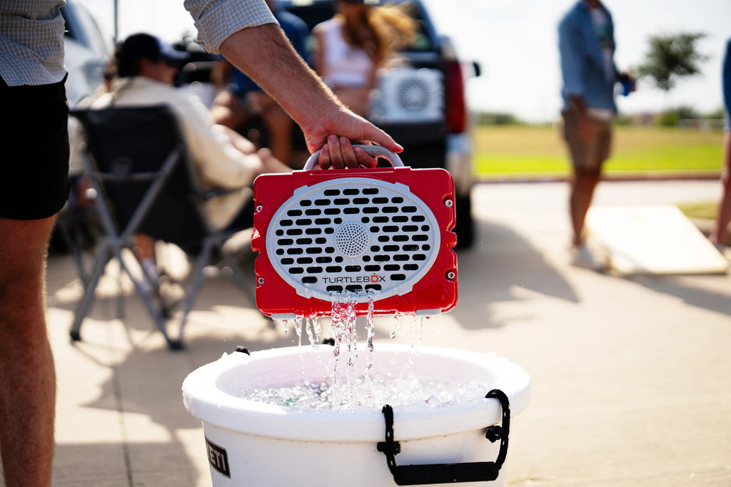 A lifestyle photo of a red and white Turtlebox portable speaker being held over a white bucket filled with water, with people and an outdoor background blurred.