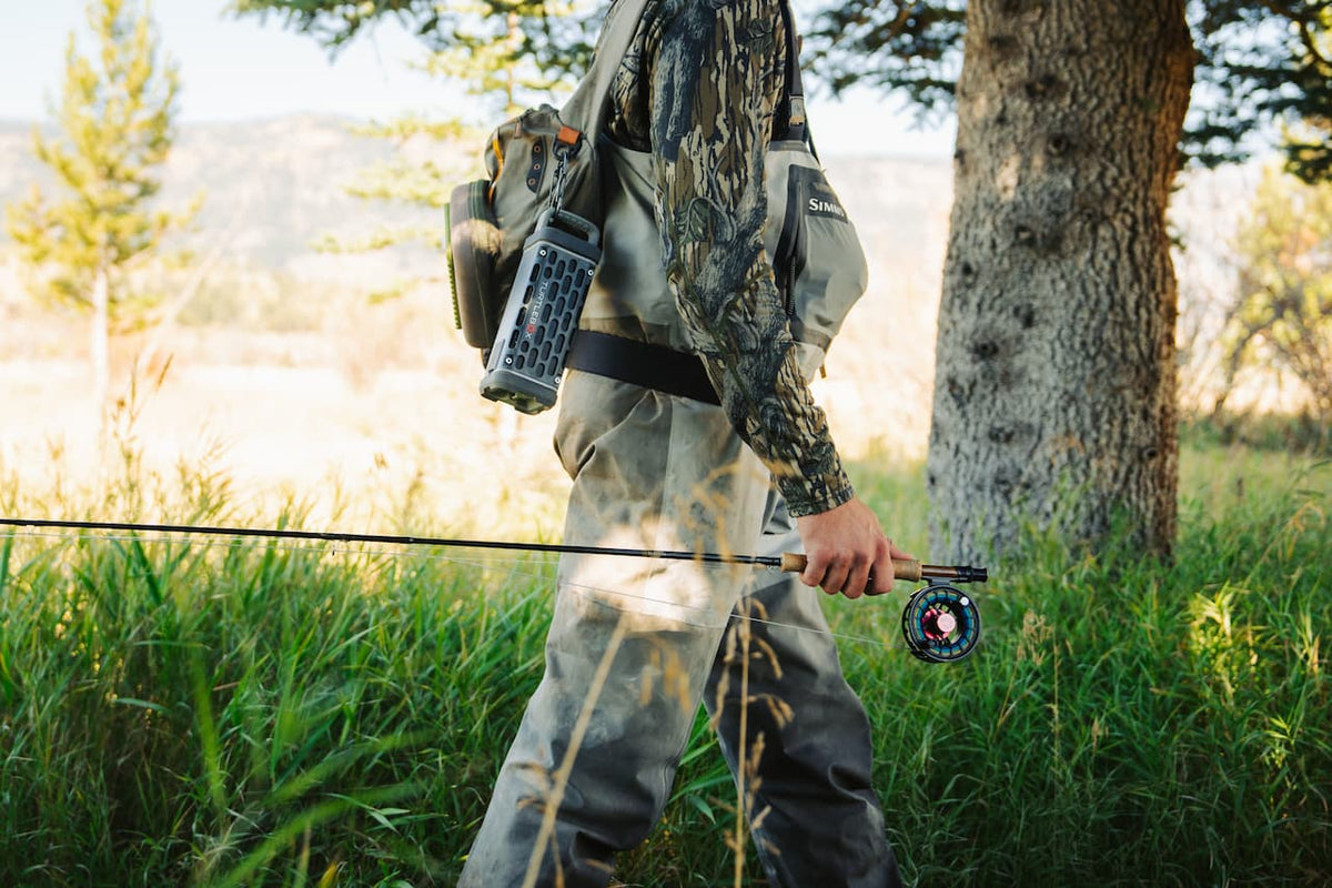 A lifestyle photo of a fisherman walking with a Ranger speaker in gray attached to his back pack. #base-color_gray