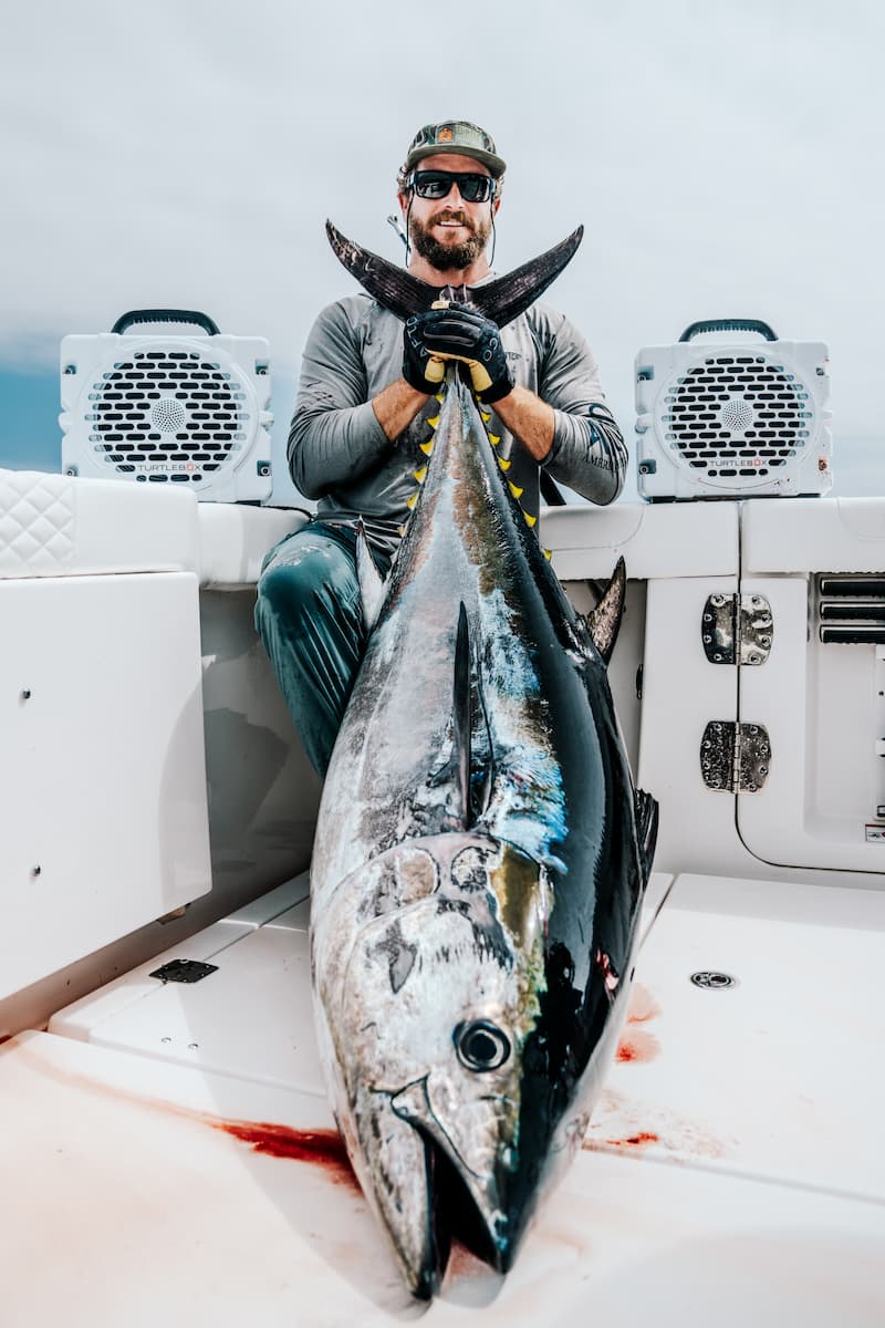 A guy in a boat holding a big tuna fish on hands with two Turtlebox Grande speakers in white color on his background
