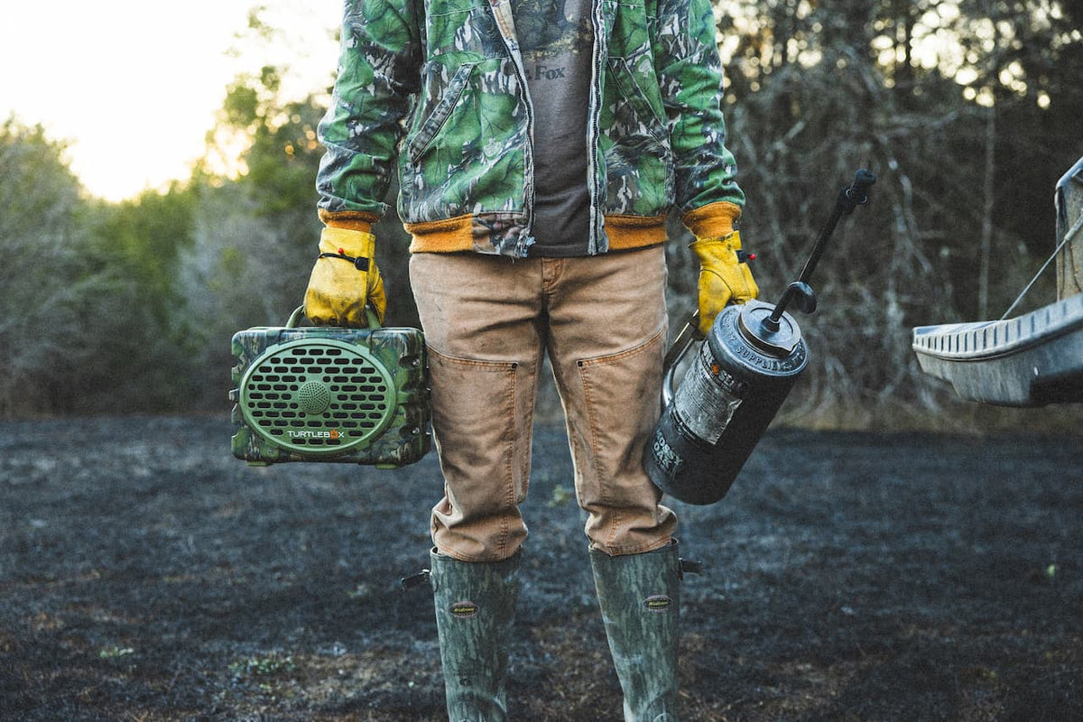 A lifestyle photo of a man holding a Turtlebox Original Mossy Oak Full Foliage edition speaker and a gasoline lighter #speaker-color_full-foliage