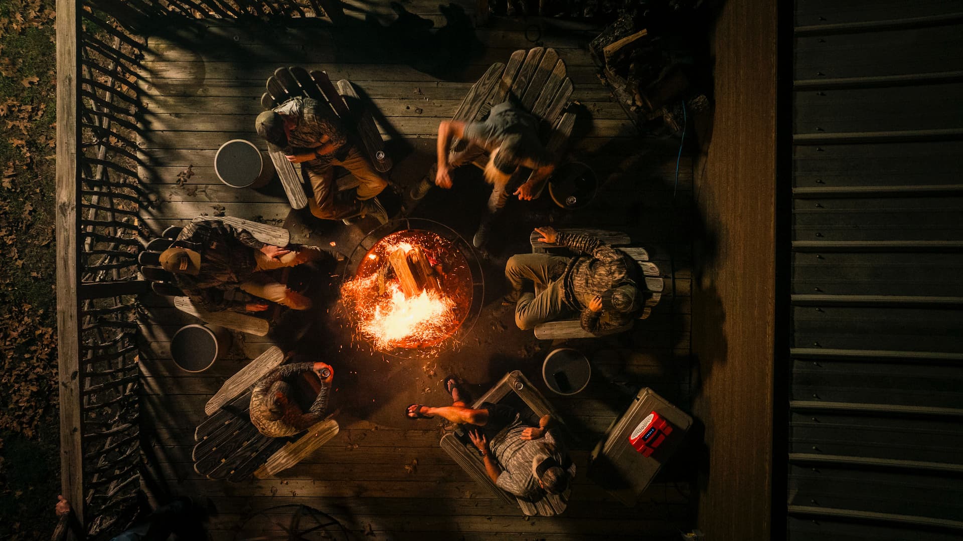 Overhead drone style shot of a group of people sitting around a campfire in adirondack chairs with an Original Orange Turtlebox speaker playing music.