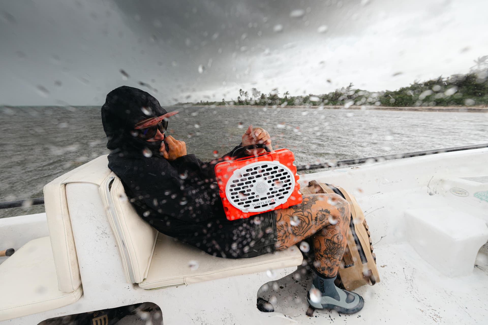 A girl sitting in a boat with a Turtlebox Original speaker in orange in a raining day