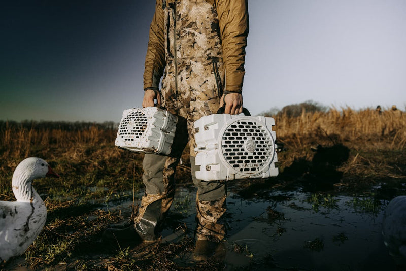 A guy in the mud with two speakers in his hands, both white color, but one Turtlebox Original speaker and the other a Turtlebox Grande speaker with some gooses walking around.