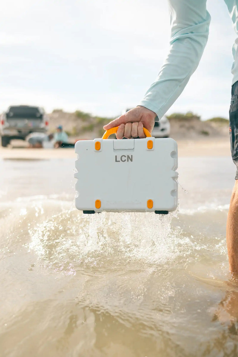 Person holding a white suitcase with orange handles on a beach