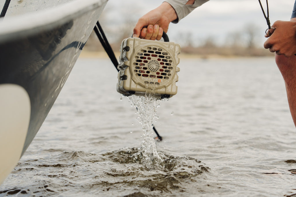 A lifestyle photo of a person holding a Turtlebox portable speaker over water, likely for cleaning or inspection purposes. #base-color_tan