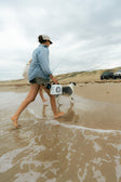 A lifestyle photo of a person walking on a beach with a dog and a surfboard, under an overcast sky. #base-color_white
