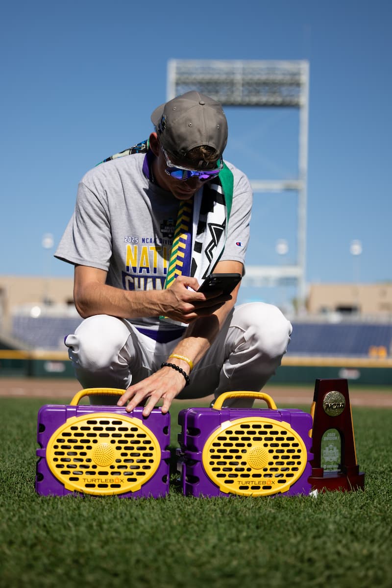 A lifestyle photo with a person using a smartphone on a purple and yellow turtlebox on a sports field #speaker-color_purple-gold-gameday-series