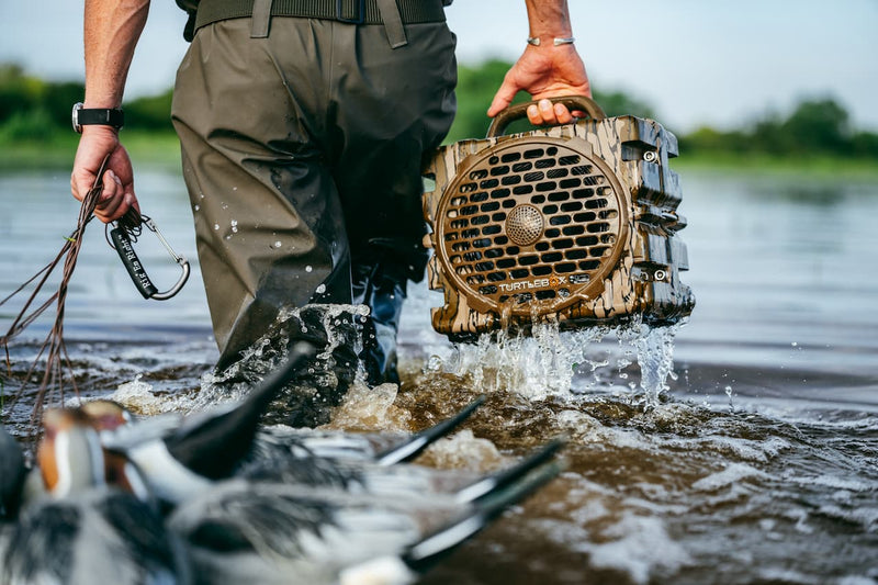 A person wading through shallow water while carrying a rugged, water-resistant wooden speaker filled with duck calls. The speaker is shown splashing water as it is lifted from the water, with ducks visible in the foreground.