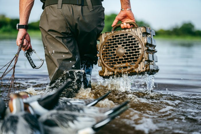 A person wading through shallow water while carrying a rugged, water-resistant wooden speaker filled with duck calls. The speaker is shown splashing water as it is lifted from the water, with ducks visible in the foreground.