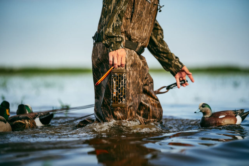 A duck hunter in full camouflage stands in shallow water holding a live bird and a release device, with mallards swimming around in a natural lake setting.