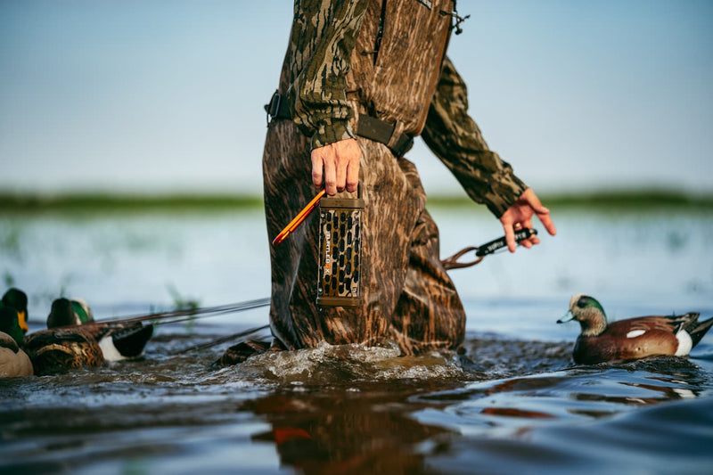 A duck hunter in full camouflage stands in shallow water holding a live bird and a release device, with mallards swimming around in a natural lake setting.