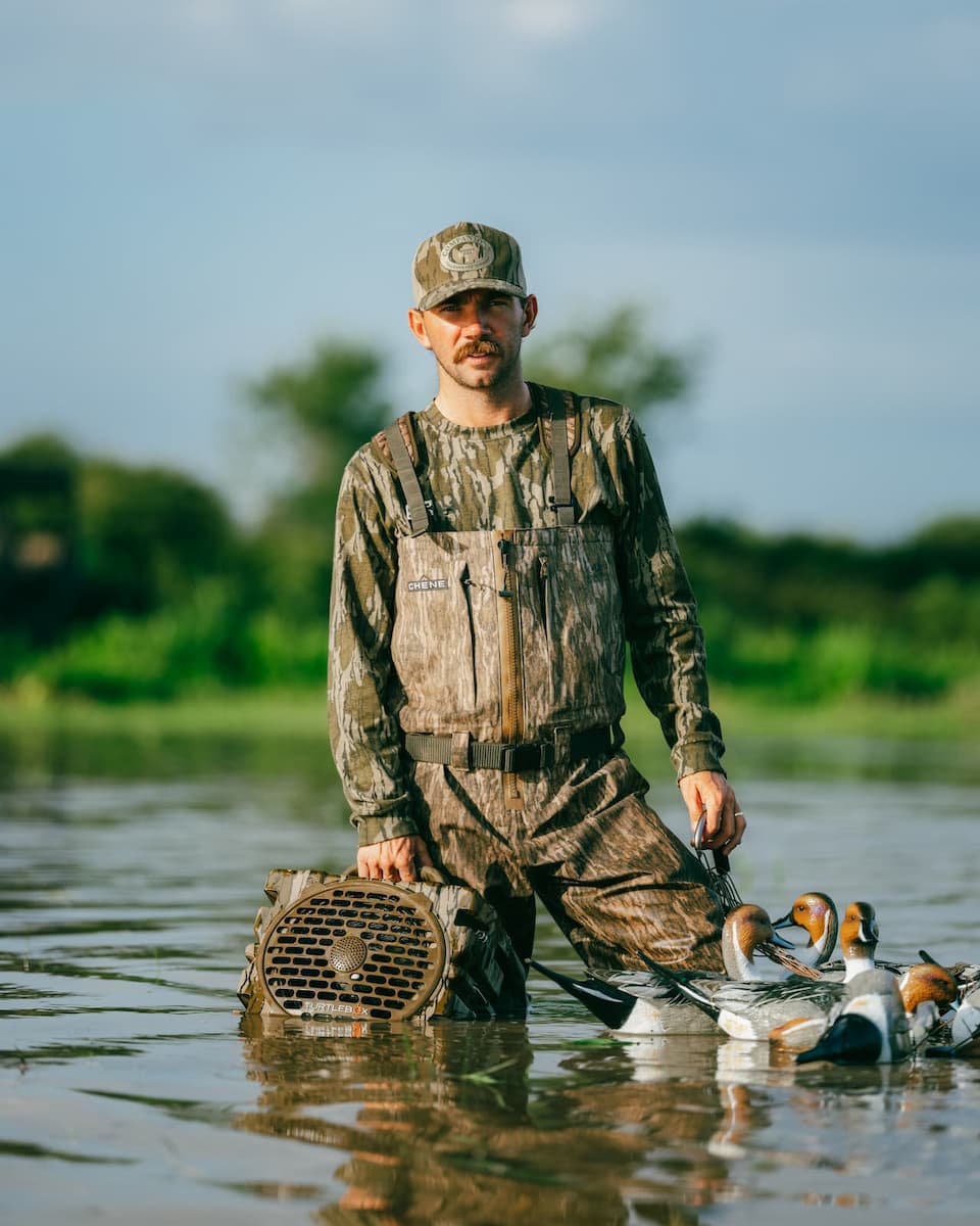 A man in full camouflage duck hunting overalls stands knee-deep in a lake holding a duck deco, with several ducks floating on the water in front of him under a blue sky.