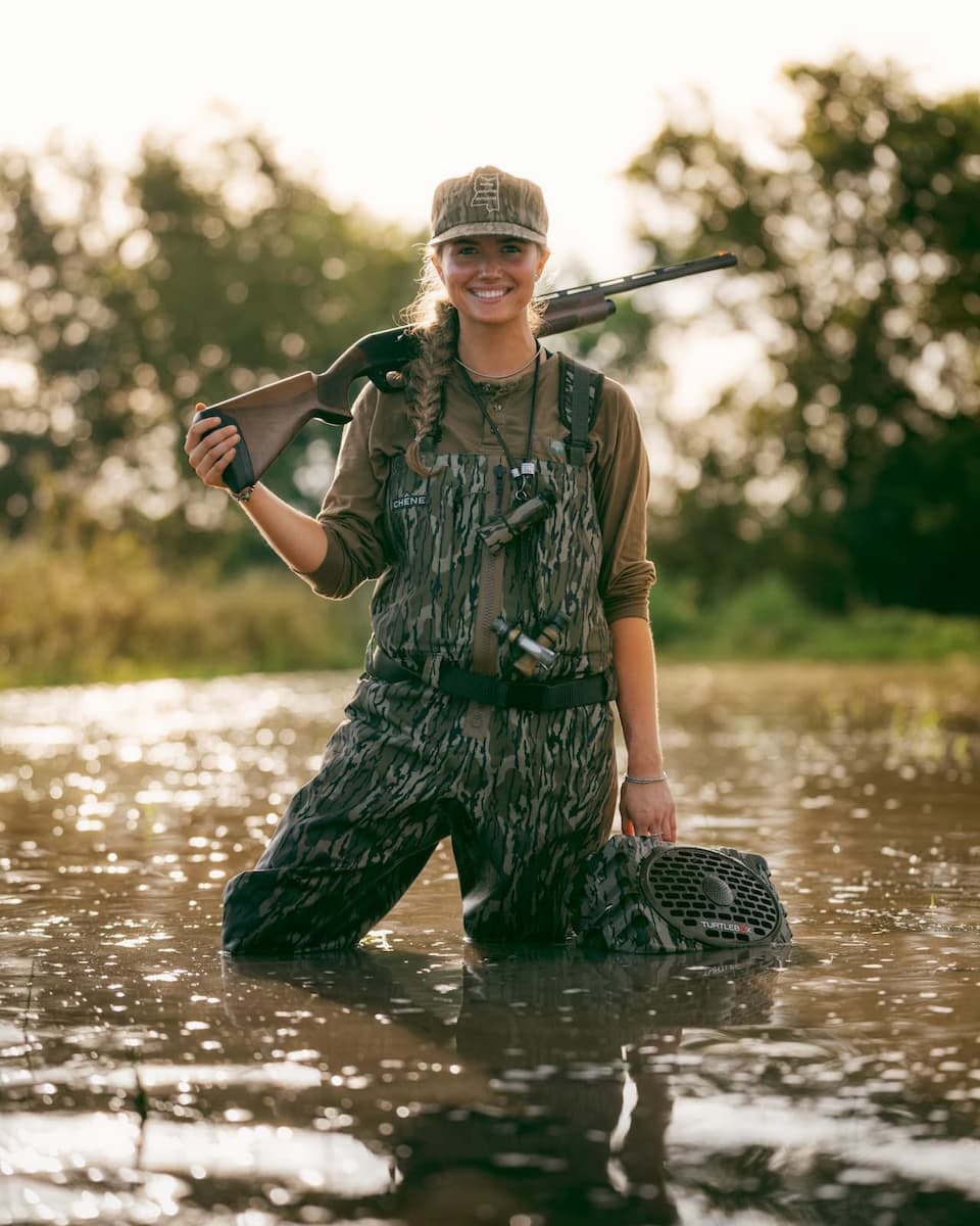 A woman in camouflage hunting overalls stands knee-deep in a river, holding a shotgun over her shoulder and a landing net in the water, with trees blurred in the background.