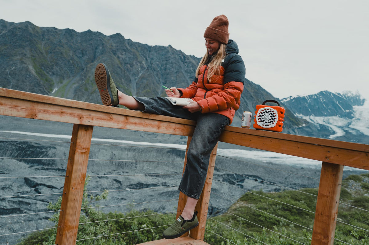 A woman sits on a wooden railing overlooking a mountain and glacier landscape, wearing a rust-brown beanie, a black and orange puffer jacket, dark jeans, and green hiking shoes. A small orange portable radio sits on the railing beside her.
