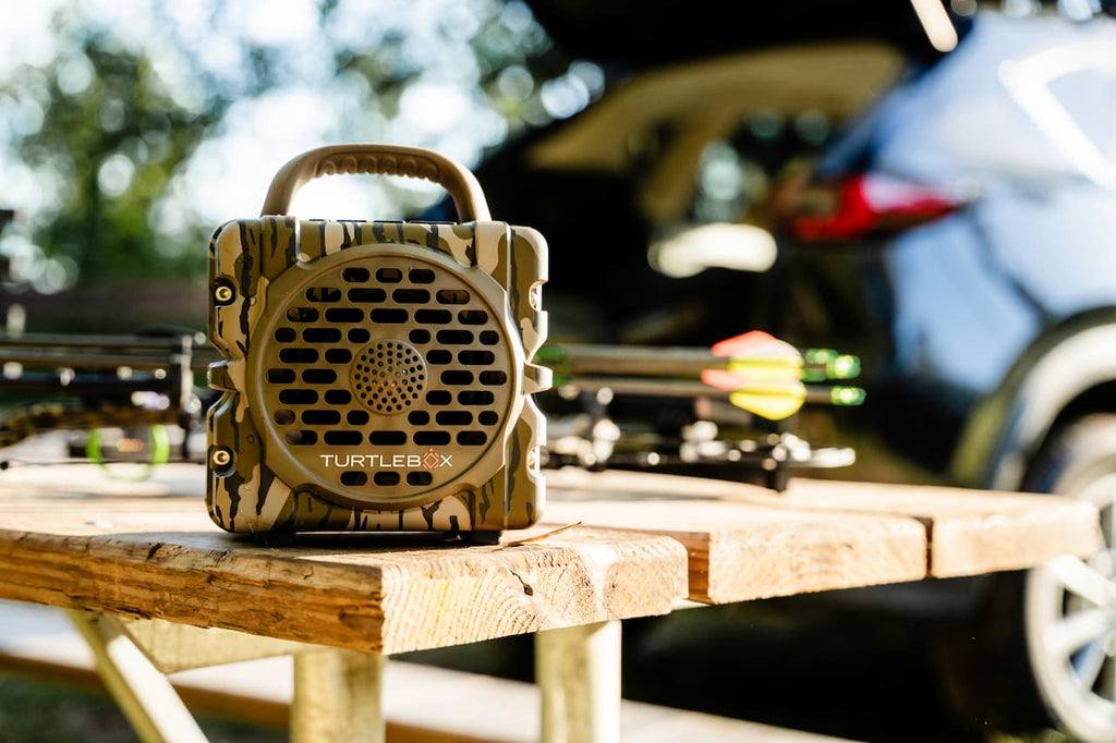 A TurtleBix portable speaker with a camouflage pattern sits on a wooden table outdoors, with archery equipment and a blurred car in the background.