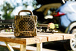 A TurtleBix portable speaker with a camouflage pattern sits on a wooden table outdoors, with archery equipment and a blurred car in the background.