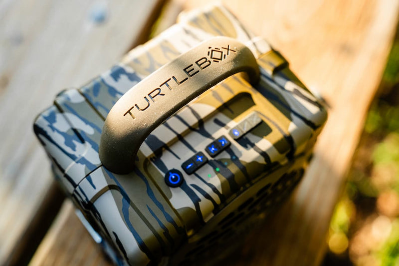 A close-up shot of a rugged, camouflage-patterned TurtleBox portable speaker resting on a wooden surface outdoors.