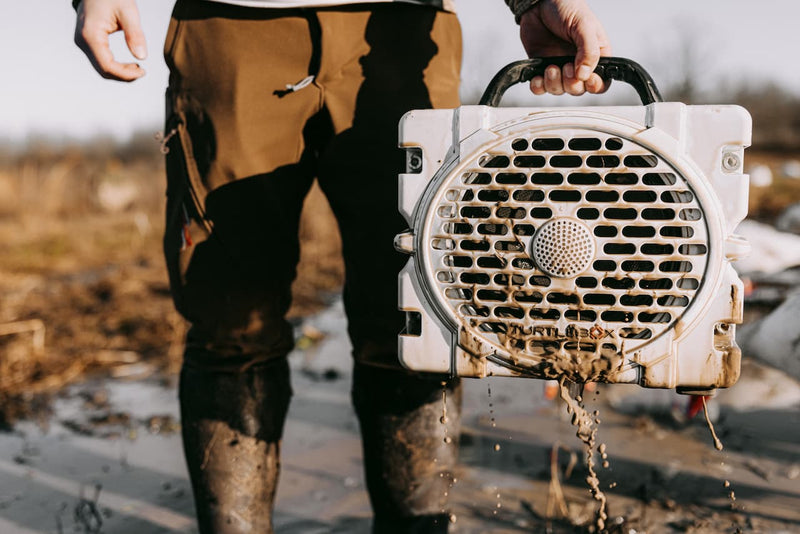 A lifestyle photo of a man in mud holding a Turtlebox Grande speaker in white color #speaker-color_white