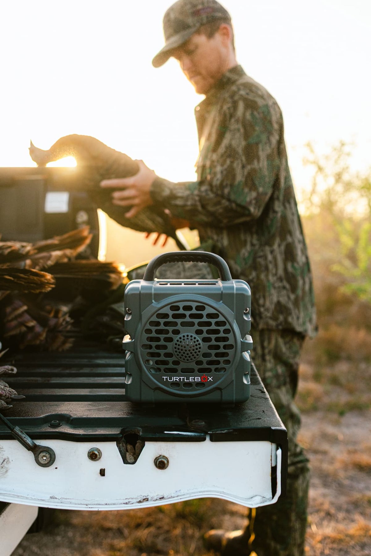 Person in camouflage holding a deer next to a Turtleback hunting backpack on a vehicle, with a sunset background.