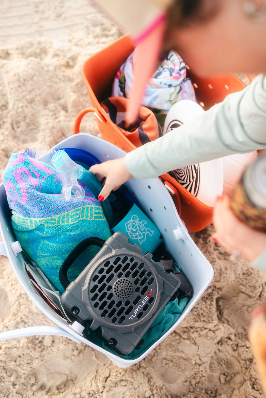 A lifestyle photo of a hand reaching into a bag with a Cub Gray speaker and beach items on sand #base-color_gray