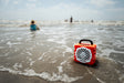 A lifestyle photo of an orange portable radio on a beach with people in the background #base-color_orange