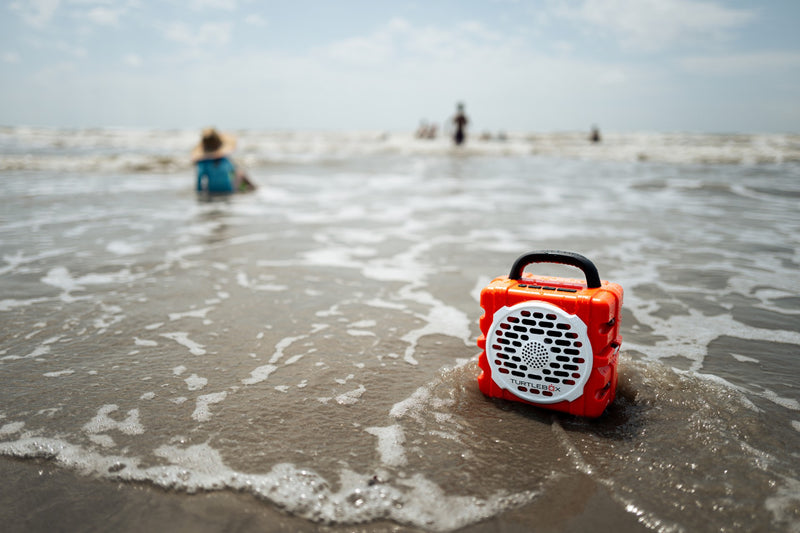 A lifestyle photo of an orange portable radio on a beach with people in the background #base-color_orange