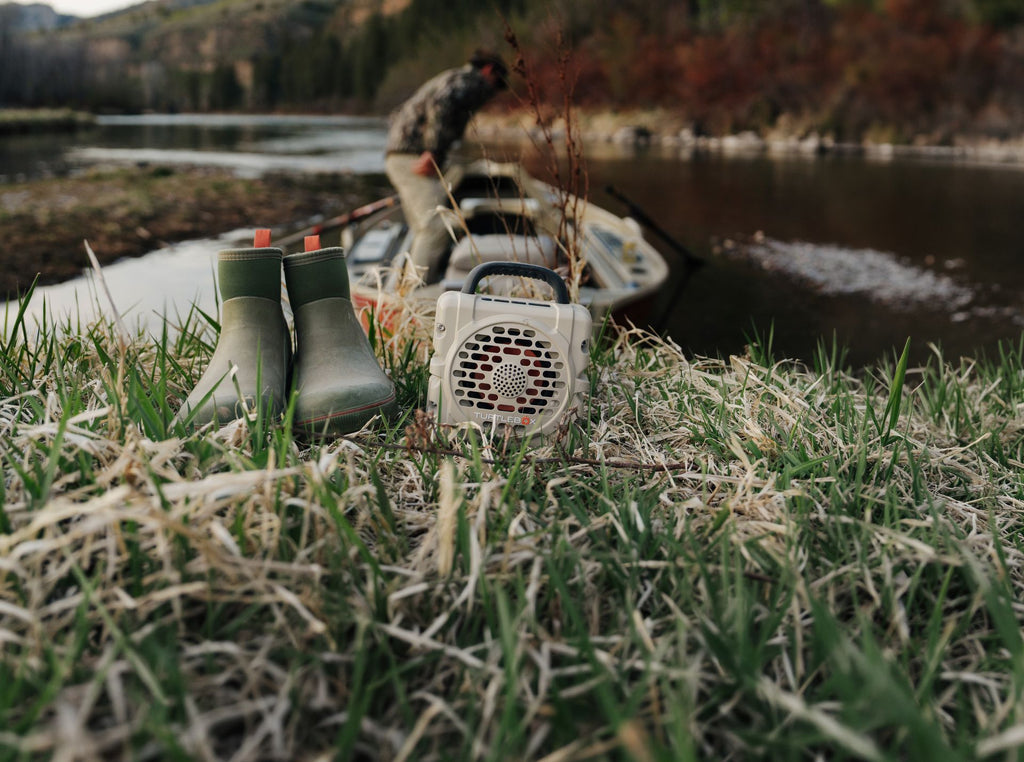 A lifestyle photo of a green rubber boots and a small Turtlebox portable speaker on grass near a body of water. #base-color_tan