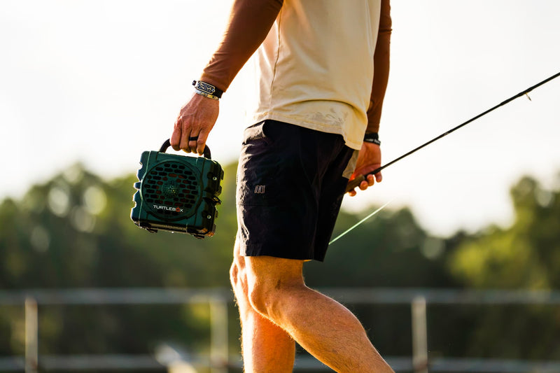 A man outdoors carrying a green and black portable speaker in one hand while holding a fishing rod in the other, wearing a cream t-shirt and black shorts. #base-color_green
