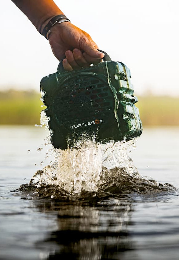 A hand holding a green waterproof speaker above splashing water outdoors, with a blurred natural background.