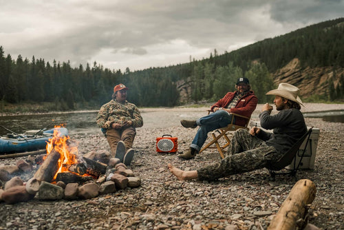 Three people sitting around a campfire by a river with a forest in the background