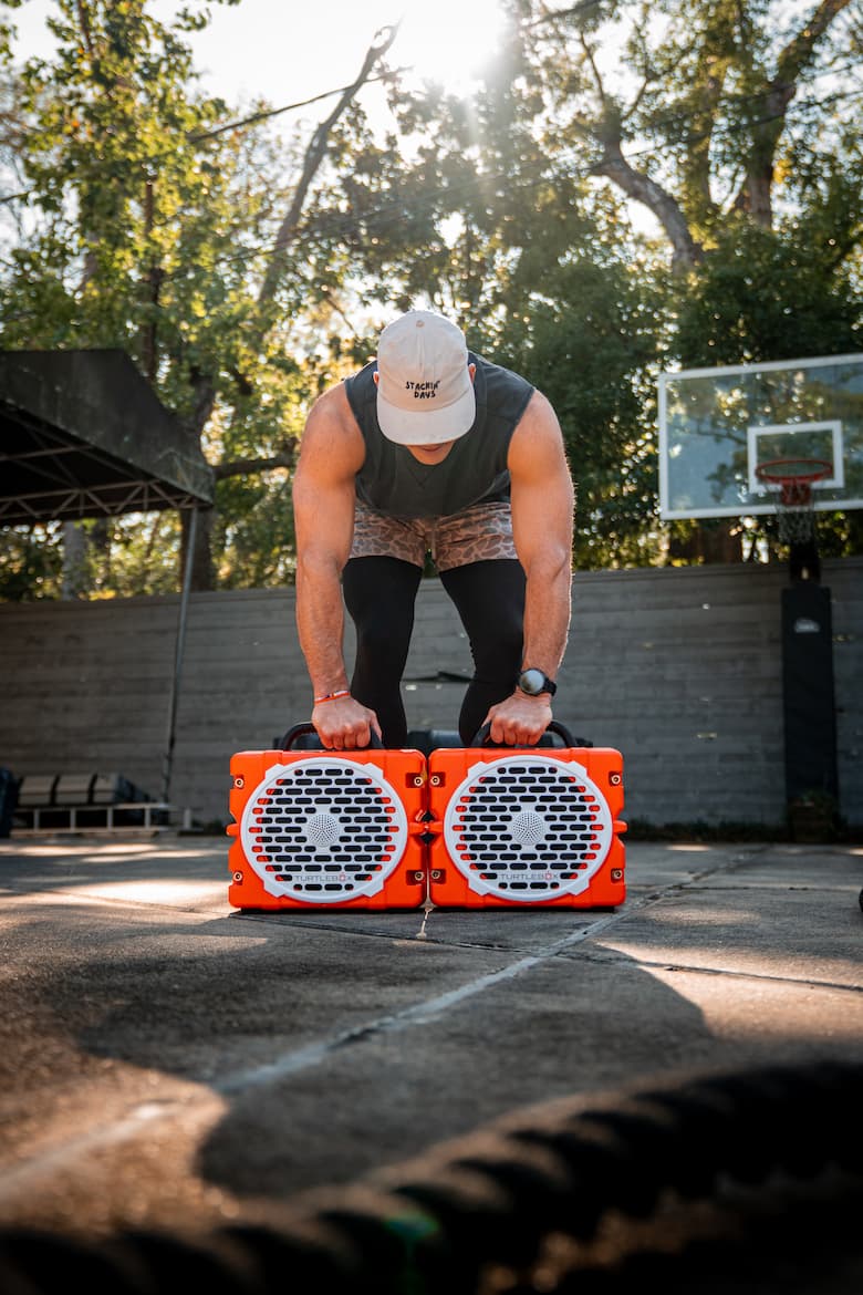 A man in athletic wear bends forward over two bright orange portable speakers on an outdoor basketball court, with trees and a basketball hoop in the background.