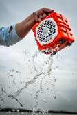 A lifestyle photo of an orange and white portable speaker being held underwater with water splashing around it. #base-color_orange
