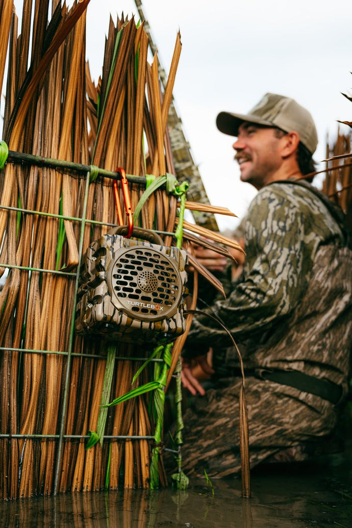 A man in camouflage clothing and a cap sits in shallow water beside a large reed or bamboo wall structure, with a small camouflage speaker or trap device hanging on the wall.