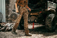 A person wearing camouflage hunting pants and a brown shirt walks beside a rugged off-road vehicle, holding a cooling fan. Ducks and a brick wall are visible in the outdoor setting.