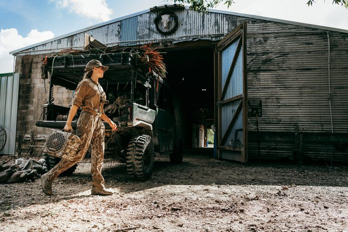 A woman in camouflage hunting clothing walks out of a rustic corrugated metal barn toward a camouflage hunting vehicle parked inside. She is holding a shotgun.