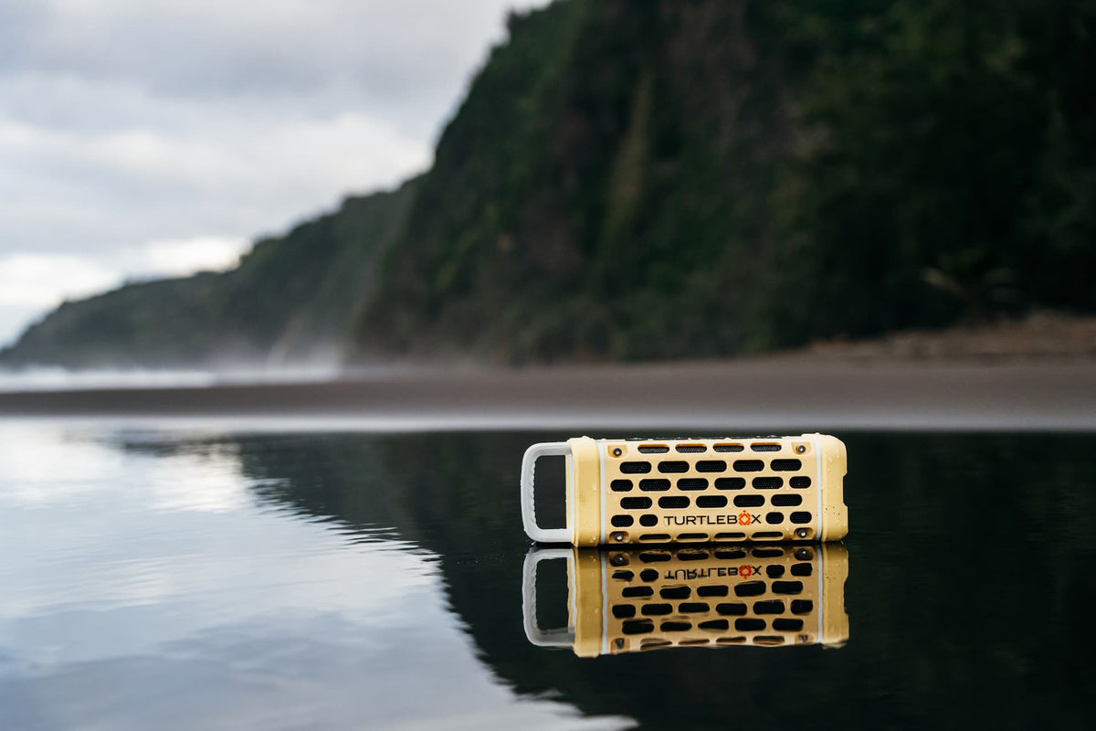 A lifestyle photo of a Fighting Lady Yellow Turtlebox Ranger speaker on a reflective water surface with a mountainous background #base-color_fighting-lady-yellow