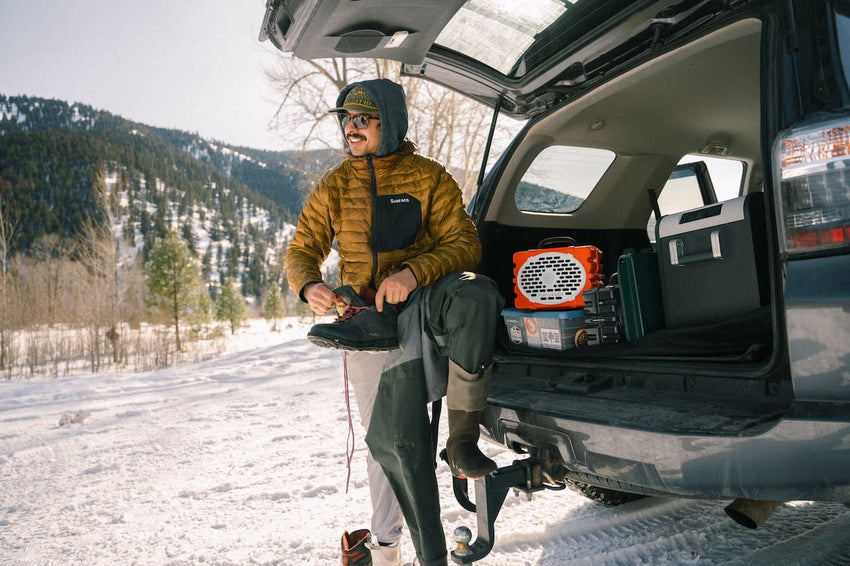 Person preparing for a winter activity with equipment in an open car trunk, snowy landscape.