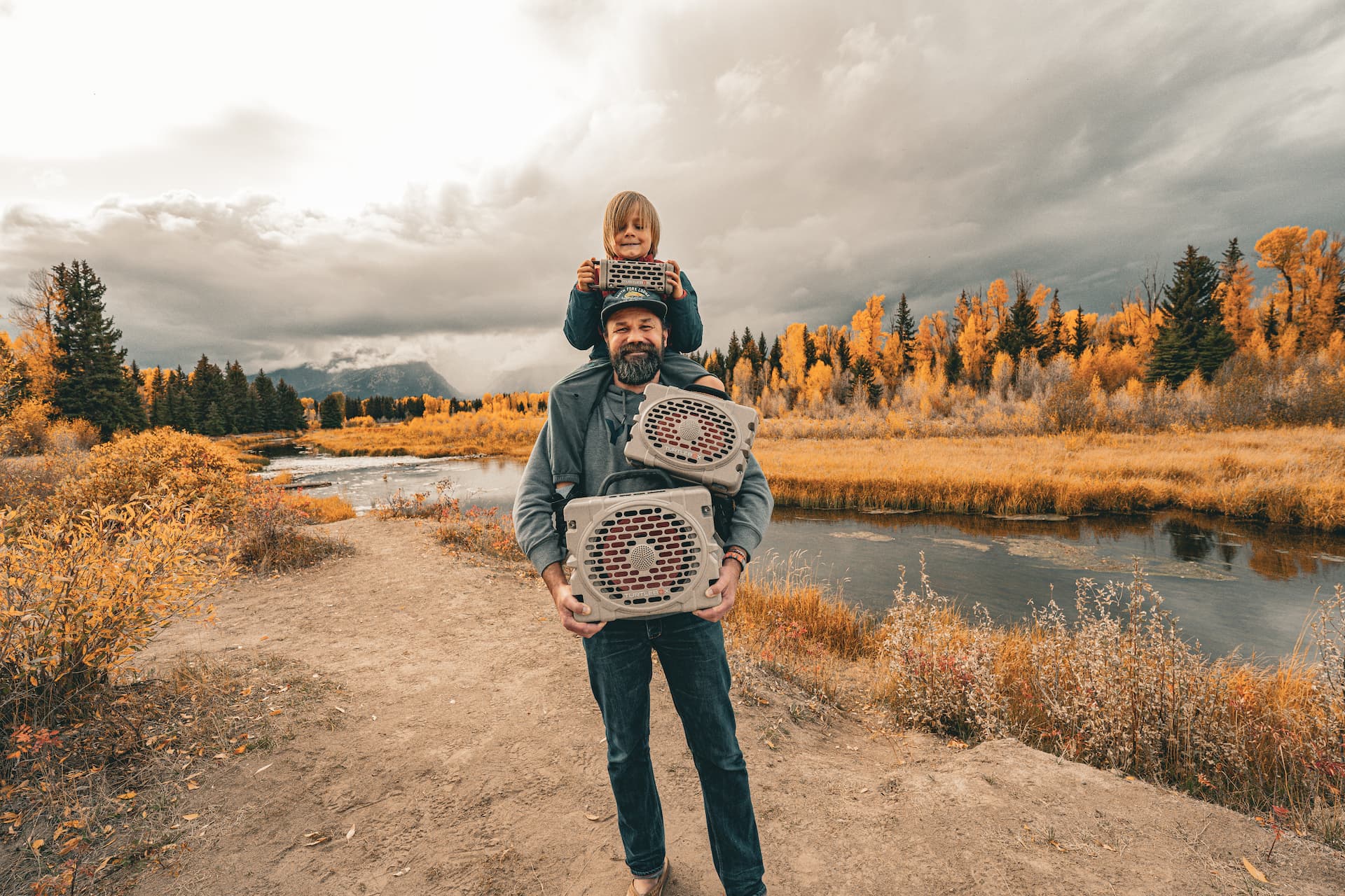 Man holding a child on his shoulders with Turtlebox speakers in a scenic outdoor setting.