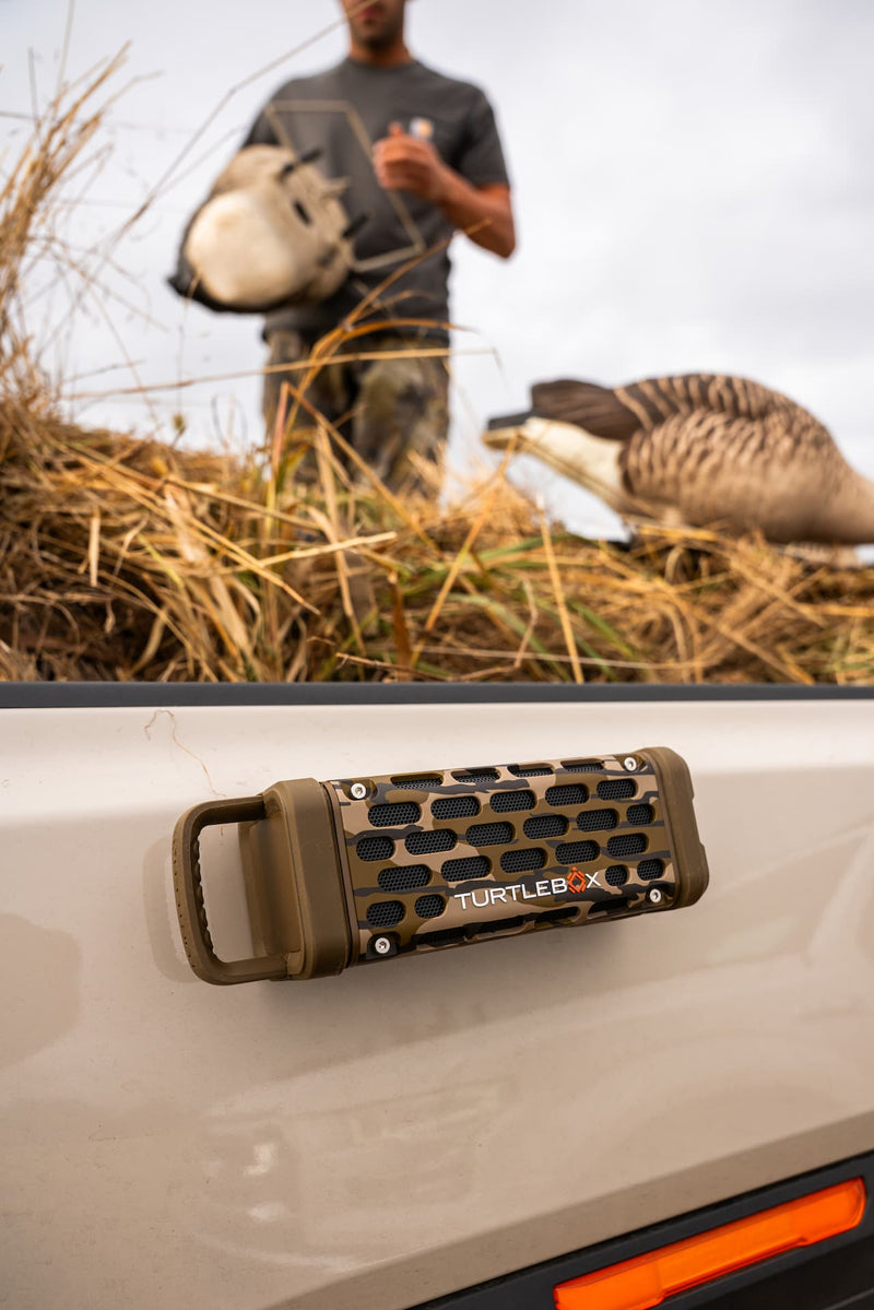 A close-up of a TURTLEBOX X portable speaker mounted on the side of a vehicle, with a blurred outdoor hunting scene in the background showing a person and a goose.